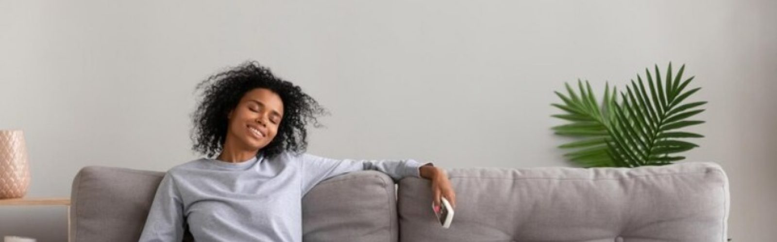 Woman relaxing on a gray sofa with a remote, enjoying comfort in a well-cooled room, indoor plants in the background, emphasizing the benefits of ductless air conditioning.